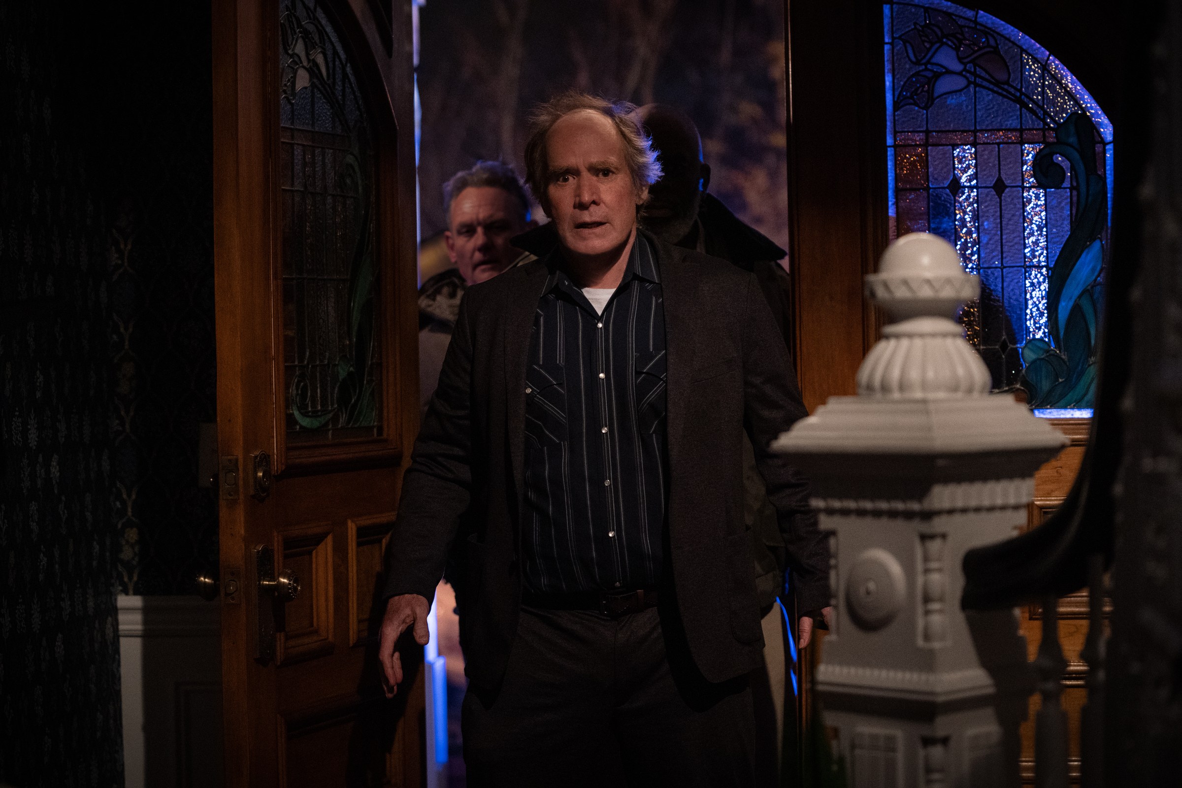 Hawkins emerges from the house beside a stained-glass door.