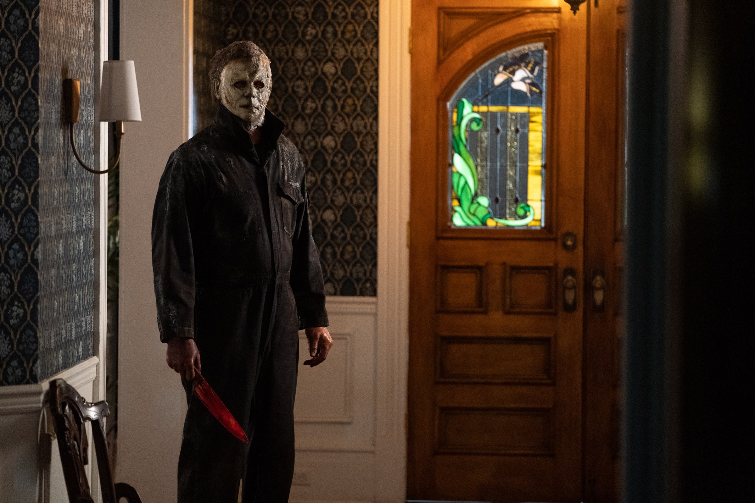 The masked killer stands beside the stained-glass front door.