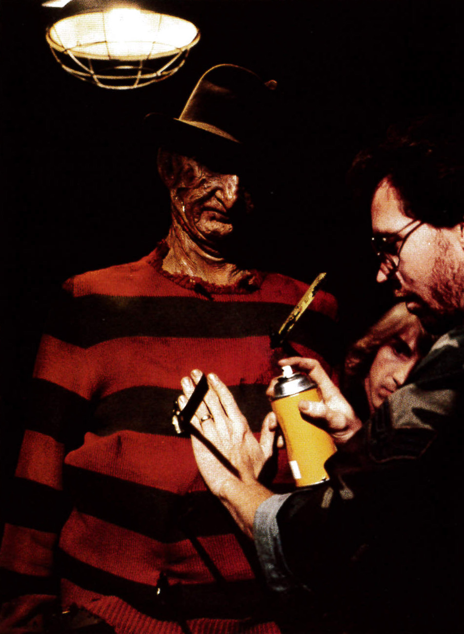 Robert Englund under a work light while an artist adjusts his makeup.