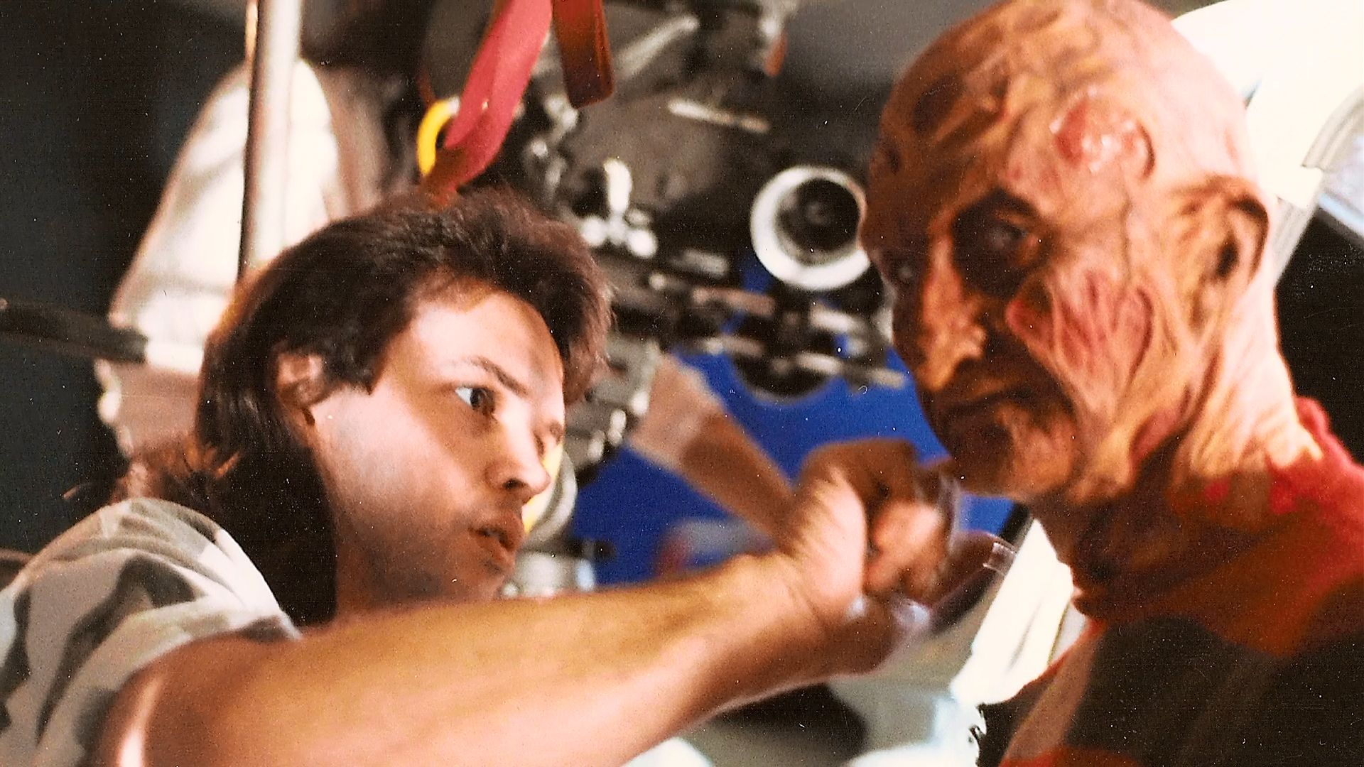 An artist retouches Robert Englund's face with camera equipment visible behind them.